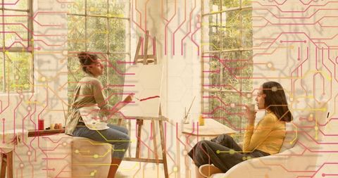 Artist painting bold red stroke on canvas in sunlit studio while companion sipping tea and chatting