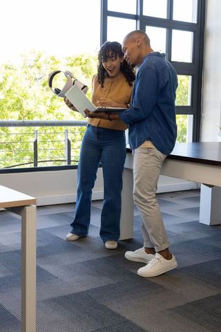 Diverse coworkers standing, discussing laptop with headphones in bright modern workspace