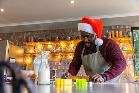 African american man baking christmas cookies wearing santa hat in cozy rustic kitchen