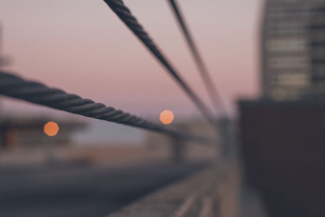 This stock photo showcases a blurred cityscape at dusk with metal cables in the foreground. The soft colors and bokeh effect create a serene, abstract urban scene. Suitable for use in projects related to city life, twilight environments, and abstract backgrounds.