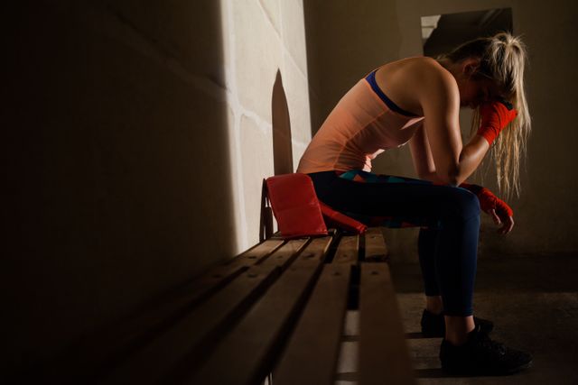 Woman in athletic wear sitting on a bench in a fitness studio, appearing sad and contemplative. This image can be used to depict themes of mental health, emotional stress, and the challenges faced during workouts or training sessions. Suitable for articles, blogs, or campaigns related to fitness, mental well-being, and personal struggles.