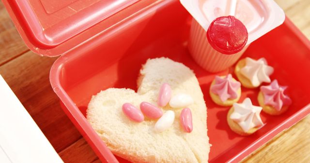 Heart-shaped sandwich, candies, small cookies, and drink placed in red lunchbox. Ideal image for Valentine's Day themes, children's lunch ideas, school lunch packing tips, snack inspirations. Perfect for blog posts, food magazines, lifestyle articles, and promotional materials for lunch products or pastry shops.