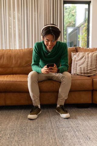 Asian man wearing headphones texting on smartphone while sitting on brown leather sofa