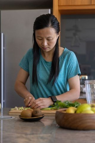 Middle-aged asian woman chopping green vegetables on kitchen island wearing smartwatch