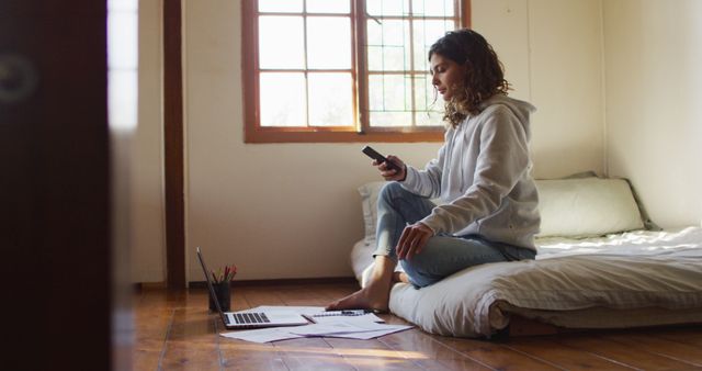 Biracial woman working at home, sitting on bed using smartphone and laptop in cottage - Download Free Stock Photos Pikwizard.com
