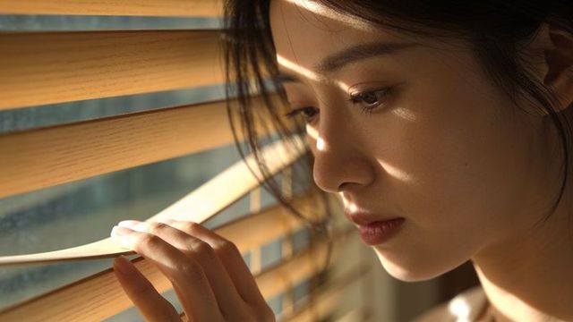 Sunlit contemplative asian woman peering through blinds closeup profile morning light