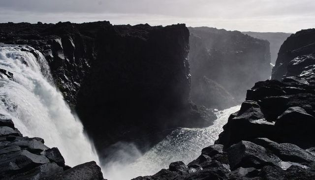 Plunging waterfall carving jagged basalt canyon under moody overcast sky, mist rising