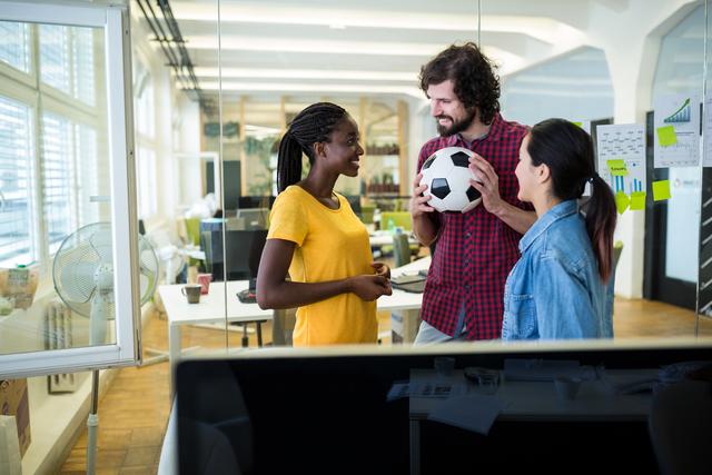 Creative team of graphic designers interacting in a modern office. They are engaged in a casual conversation, holding a soccer ball, indicating a friendly and collaborative work environment. This image can be used to depict teamwork, office culture, and a positive work atmosphere in marketing materials, websites, and presentations.