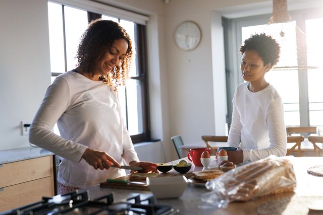 Happy, diverse lesbian couple preparing breakfast standing in sunny kitchen. Togetherness, free time and domestic life concept.