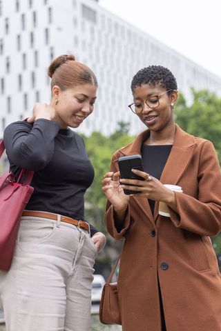 African american women checking smartphone together on city street holding coffee and tote