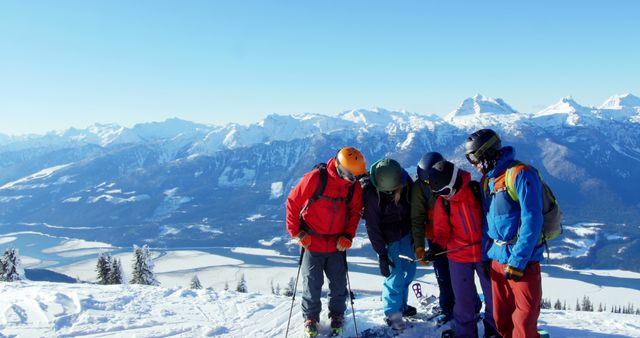 Four skiers in bright jackets and helmets examining their equipment while standing on a snowy mountain. The mountain range offers a breathtaking, panoramic view with clear blue skies in the background. Ideal for use in articles or advertisements focusing on winter sports, active lifestyle, outdoor adventure, or travel and tourism in alpine regions.