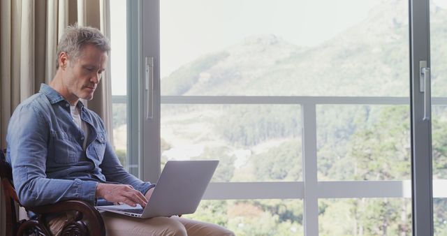 Older man sitting by a window with mountain view using laptop. Perfect for depicting remote work environments, home office setups, tranquility during work, or senior professionals adapting to digital tools. Can be used in articles about remote work trends, relaxation while working, or technology in the lives of older adults.