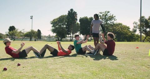 African american trainer leading teen athletes doing partner sit-ups on sunny sports field