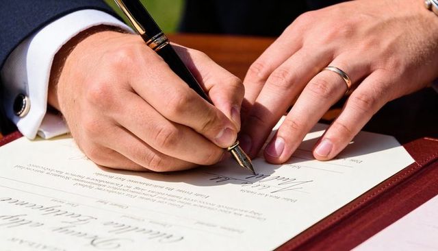 Groom signing marriage certificate with fountain pen, steadying document in leather folio