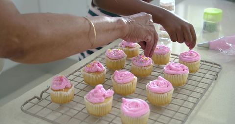 Grandmother and granddaughter decorating pink frosted cupcakes together on cooling rack