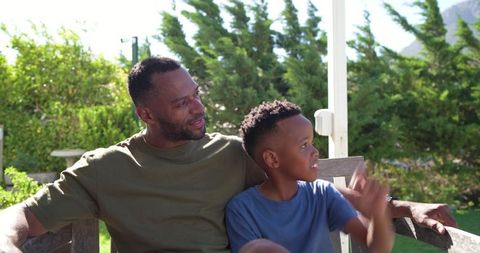 African american father and son relaxing on porch bench, child pointing toward garden