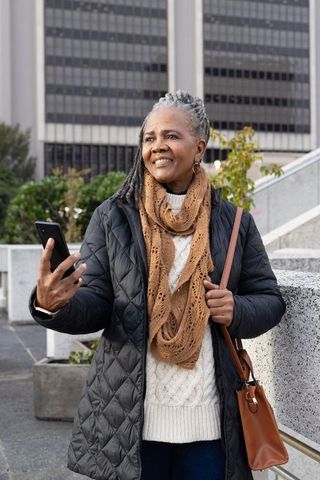 Senior african american woman smiling and holding smartphone in urban plaza wearing scarf