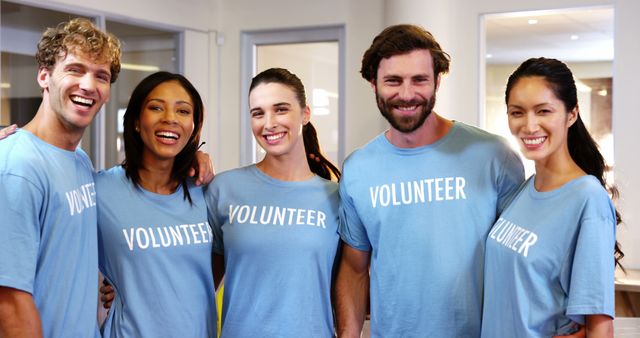Diverse group of five volunteers standing together and smiling, wearing blue volunteer shirts. Perfect for content related to community service, teamwork, charity organizations, and volunteer engagement.