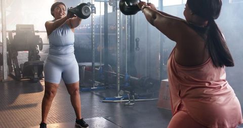 Two diverse women performing kettlebell swings facing each other partner gym workout