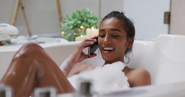 Young woman enjoying a bubble bath while talking on phone. She is smiling and appears to be having a pleasant conversation. Candles and green plants near the bath create a spa-like atmosphere, highlighting themes of relaxation and self-care. Useful for advertisements and blog posts focused on wellness, self-care routines, relaxation products, and lifestyle topics.