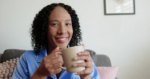 Smiling woman relaxing in blue pajamas holding ribbed beige mug on gray sofa lifestyle