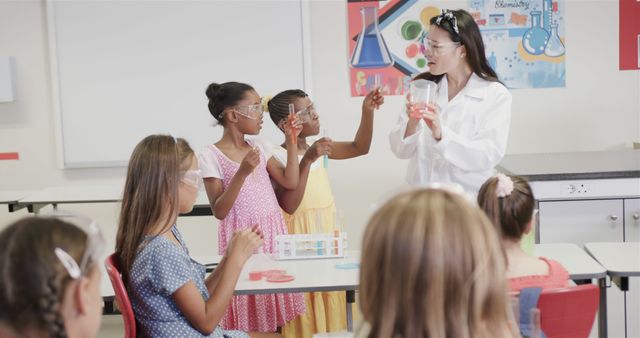 Female science teacher demonstrating a chemistry experiment to diverse group of engaged students in a primary school classroom. Students are wearing safety goggles and participating eagerly. Perfect for educational content, classroom technology, STEM education promotion, and science-related articles.