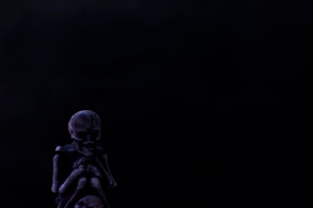 Skeleton figurine sits in dark, eerie lighting, creating spooky and Halloween-themed atmosphere. Useful for Halloween decorations, horror themes, and ghostly moods.
