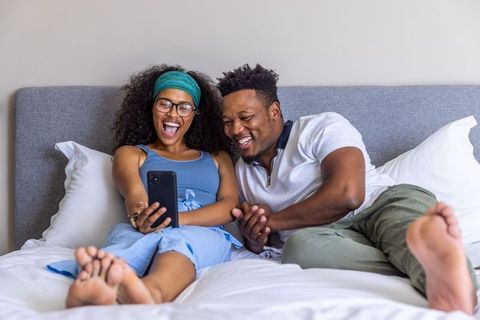 Black couple laughing on bed sharing smartphone moments for cozy at-home romance