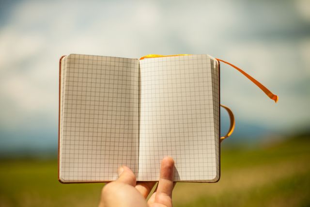 This image features a hand holding an open journal with blank grid pages against a natural outdoor background. Ideal for illustrating concepts like journaling, writing, drawing, outdoor creativity, personal goals, and organizing thoughts. Perfect for use in blogs, websites, advertisements, or educational content related to notebooks, planning, or creativity methods.