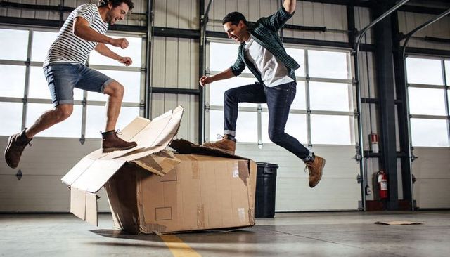 Two young men jumping over crushed cardboard box in warehouse showcasing playful energy