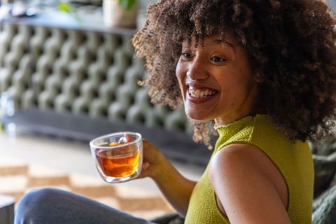 African american woman smiling while holding double-walled glass cup of tea on sofa