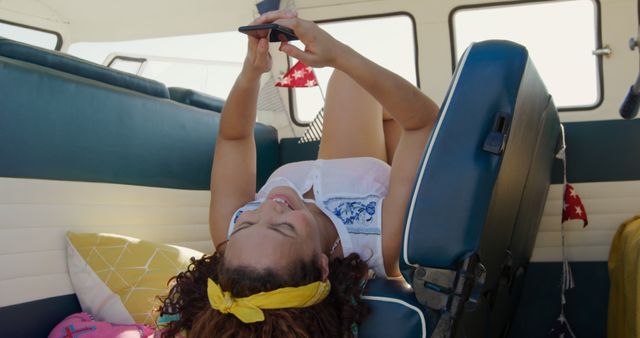 Young woman lying on a seat inside a camper van, holding and using a smartphone. Perfect for themes related to summer travel, road trips, freedom, digital nomad lifestyle, and modern leisure activities. Useful for advertisements, blogs, social media posts, and websites focused on adventure, travel, and mobile technology.