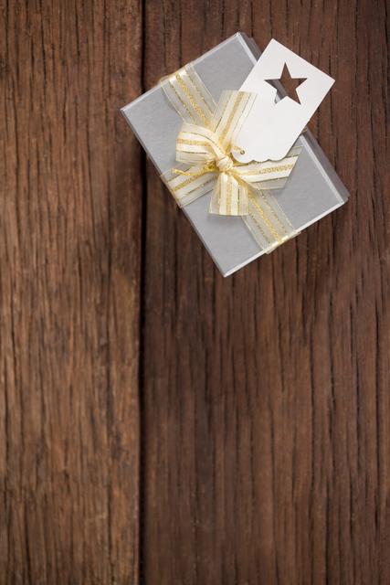 Wrapped gift box with a gold ribbon and a tag on a wooden table. Ideal for holiday promotions, Christmas advertisements, festive greeting cards, and celebration-themed designs. Perfect for illustrating gift-giving, special occasions, and festive decorations.