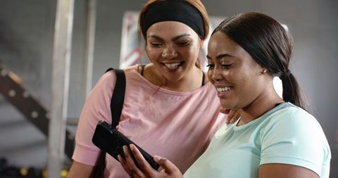 Diverse female friends smiling and checking smartphone at gym wearing activewear