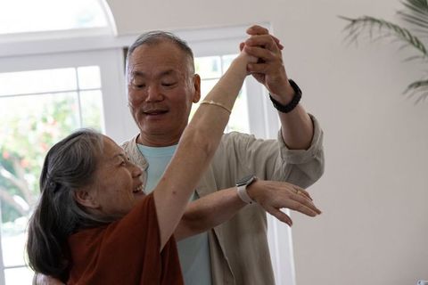 Asian senior couple dancing by sunlit window sharing joyful, affectionate at-home moment
