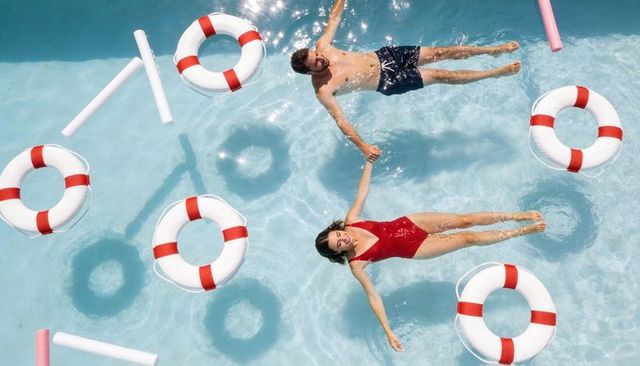 Couple floating hand-in-hand in sunny resort pool surrounded by lifebuoys and pool noodles