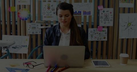 Young woman working on laptop in creative studio surrounded by sketches, color wheel