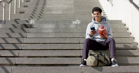 African american man sitting on concrete stairs holding football and smartphone listening