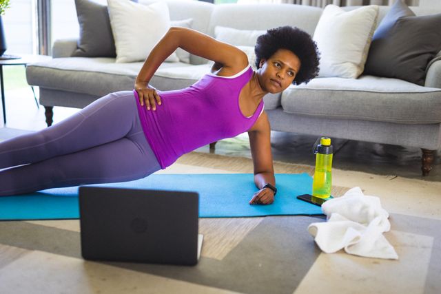 African American woman practicing yoga at home, following an online class on a laptop. She is performing a side plank on a yoga mat in her living room. This image is perfect for promoting home fitness routines, online exercise classes, healthy living, and the use of technology for personal training.