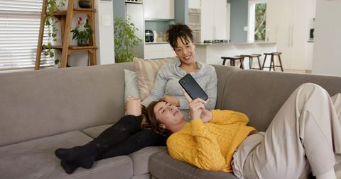 Midlife Friends Relaxing on Couch Sharing Smartphone and Coffee in Sunlit Living Room