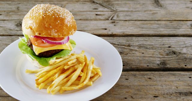 A cheeseburger with lettuce, tomato, and onion is served alongside a portion of French fries on a white plate, with copy space. The wooden table background adds a rustic touch to this classic American fast-food meal.