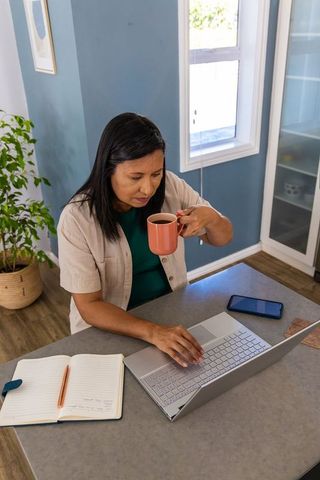 Mature woman working from home drinking coffee and using laptop at kitchen workspace
