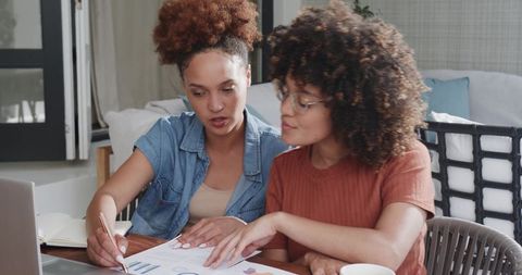 Two women collaborating on charts and reports at living room table with laptop and coffee