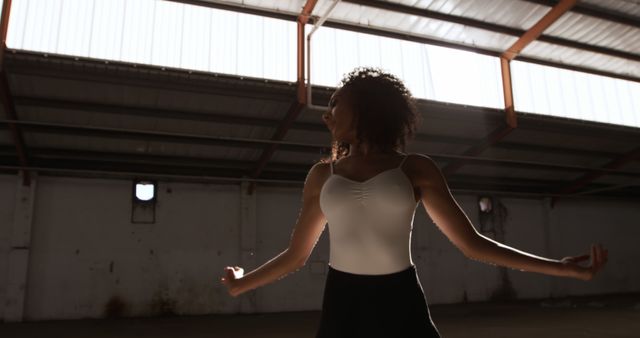 African American woman in white leotard and black skirt dancing in an industrial warehouse with natural sunlight streaming in through the ceiling. This image is perfect for use in articles, advertisements, or websites centered around dance, fitness, urban culture, or performing arts.