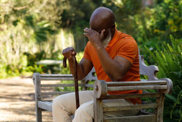 Side view of an old african-american man with a bald head and a white beard sitting on a wooden park bench. he is pressing his hand against his head while holding his cane.