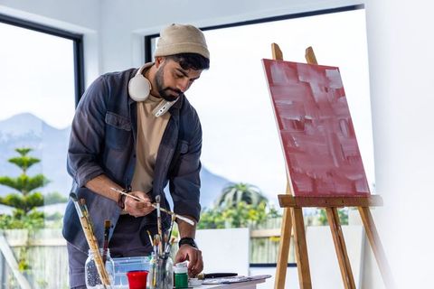 Indian artist mixing paints in bright studio with easel, brushes, palette, beanie and headphones