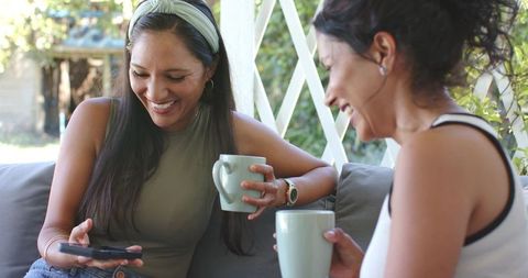 Diverse female friends laughing and sharing coffee on sunlit backyard porch sofa