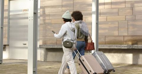Diverse female friends pulling rolling suitcases across urban plaza, casual travel