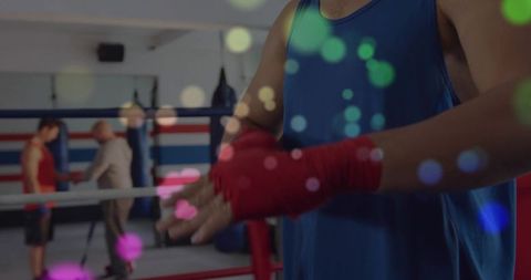 Boxer wrapping hands with red wraps in boxing ring while coach instructing, colorful bokeh