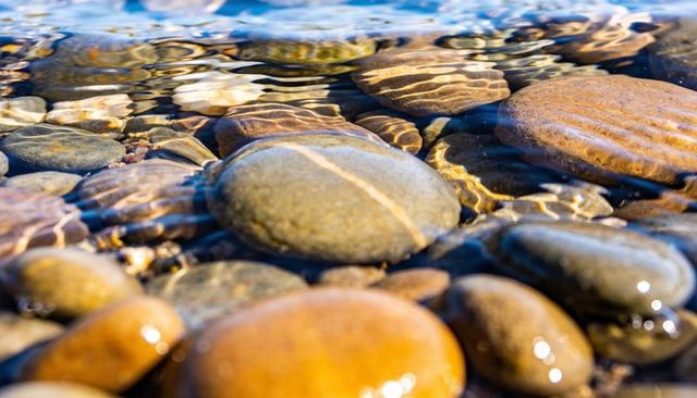 Sunlit river pebbles with water ripples and refraction bands close-up colorful texture